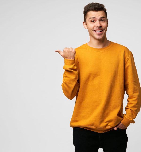 Young attractive handsome guy looks delighted gladden amazed, mouth open, smiling, like dream came true, big discount in shop,wears orange sweatshirt, points with thumb finger on a blank copyspace